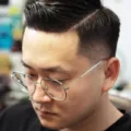 A man with short, well-groomed hair and glasses is looking down. He has a neat side part haircut and is wearing a black shirt. The background appears to be a bright indoor space with various objects, slightly out of focus.