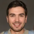 A young man with short brown hair and a beard smiles at the camera. He is wearing a light-colored shirt and a light blue backpack strap is visible over his shoulder. The background is a neutral, dark color.